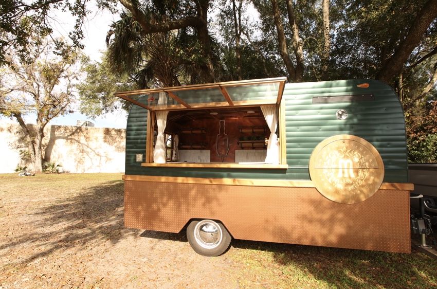 The Bar Monte trailer parked under trees: a rounded camper shape painted dark green above a copper-colored diamond-plate skirt, its glazed service hatch propped open and a large gold medallion with the Montenegro 'M' on the side.