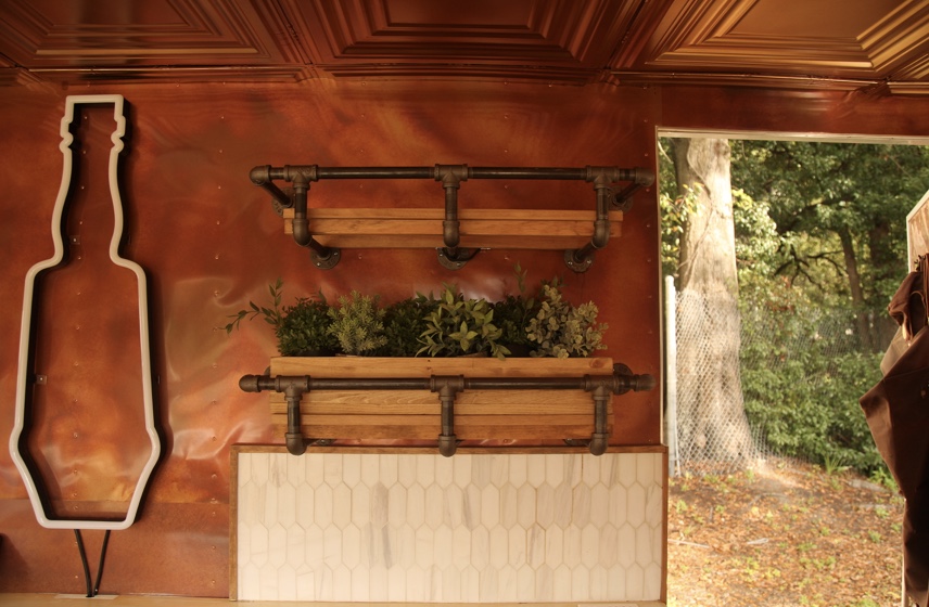 A copper-clad wall inside the Bar Monte trailer with a planter of fresh herbs on black iron pipe shelving, a tall bottle-shaped outline sign and a white tile backsplash, beside an open door to the trees outside.