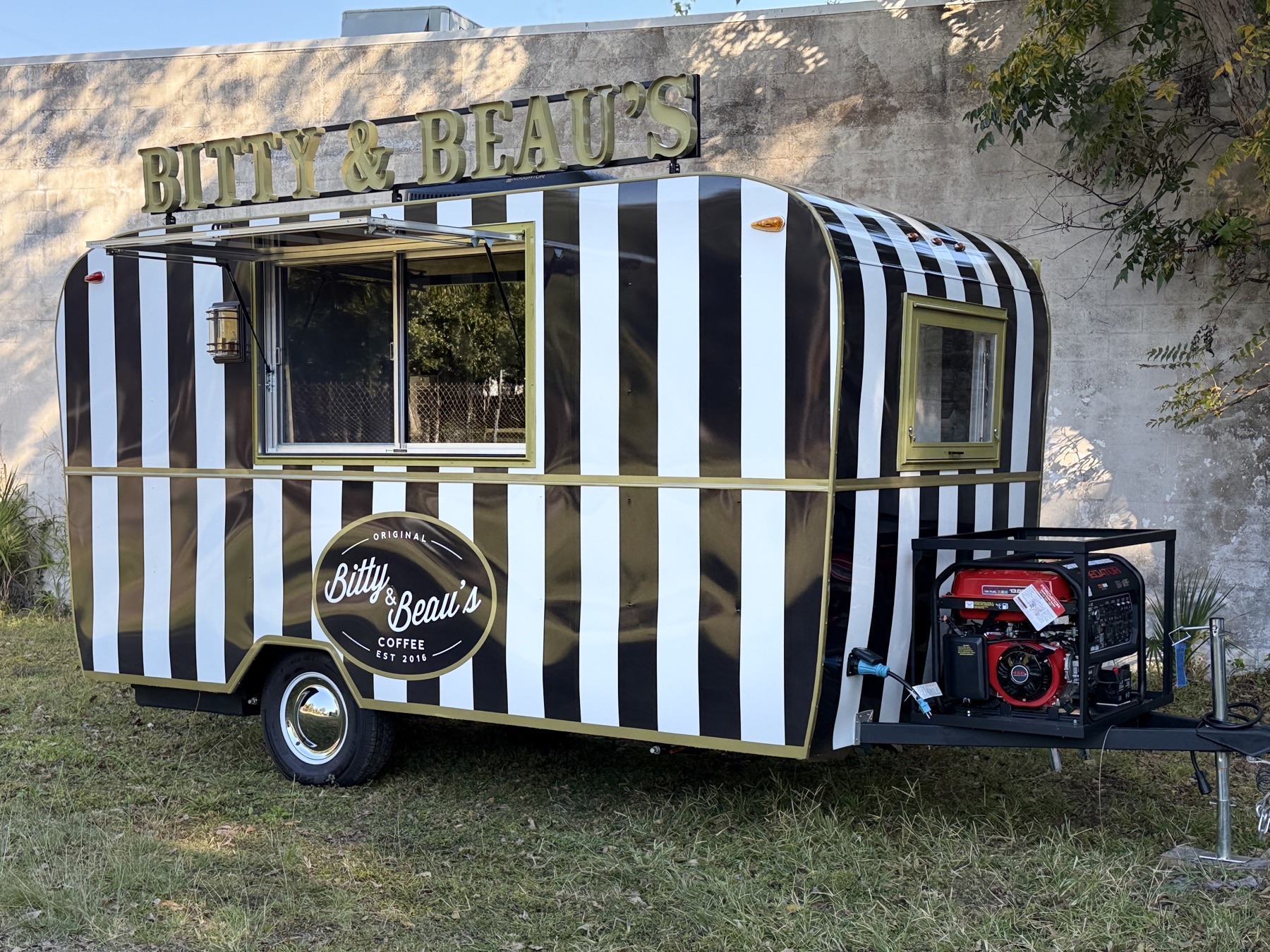 A small round-shouldered trailer in black-and-white vertical stripes with gold trim, raised gold Bitty & Beau's letters on a roof rack, an open glass service window under a hinged awning, an oval Bitty & Beau's Coffee badge on its side and a generator in a cage on the tongue.