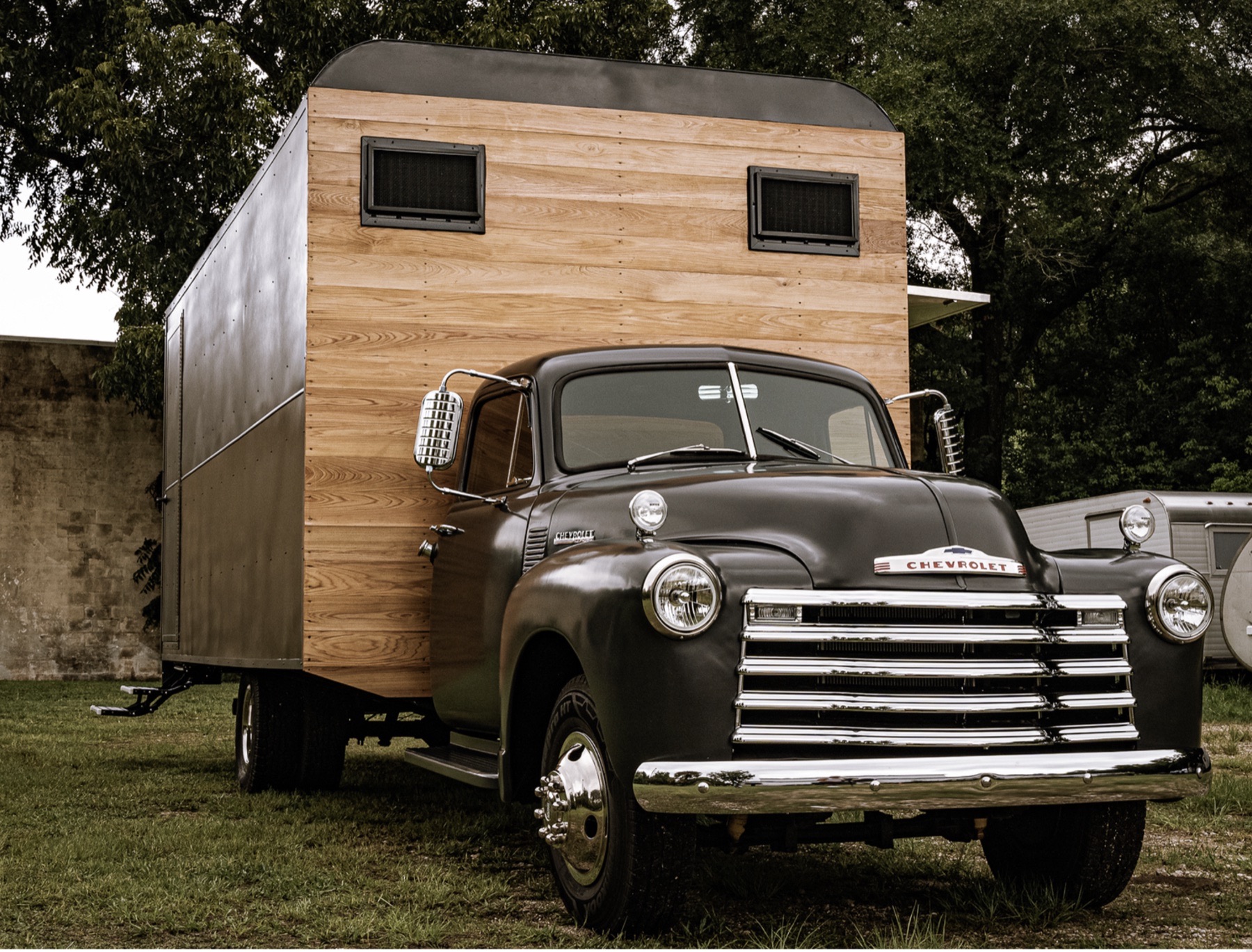 Front three-quarter view of a matte charcoal vintage Chevrolet truck with a chrome grille and Chevrolet hood badge, carrying a box body whose front wall is clad in natural wood boards.