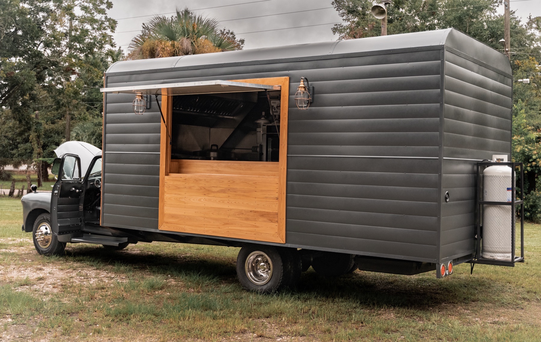 Side view of the food truck's charcoal box body with a large serving window framed in pale wood, a fold-out awning, two caged lamps, stainless kitchen equipment visible inside and a propane tank on a rear rack.