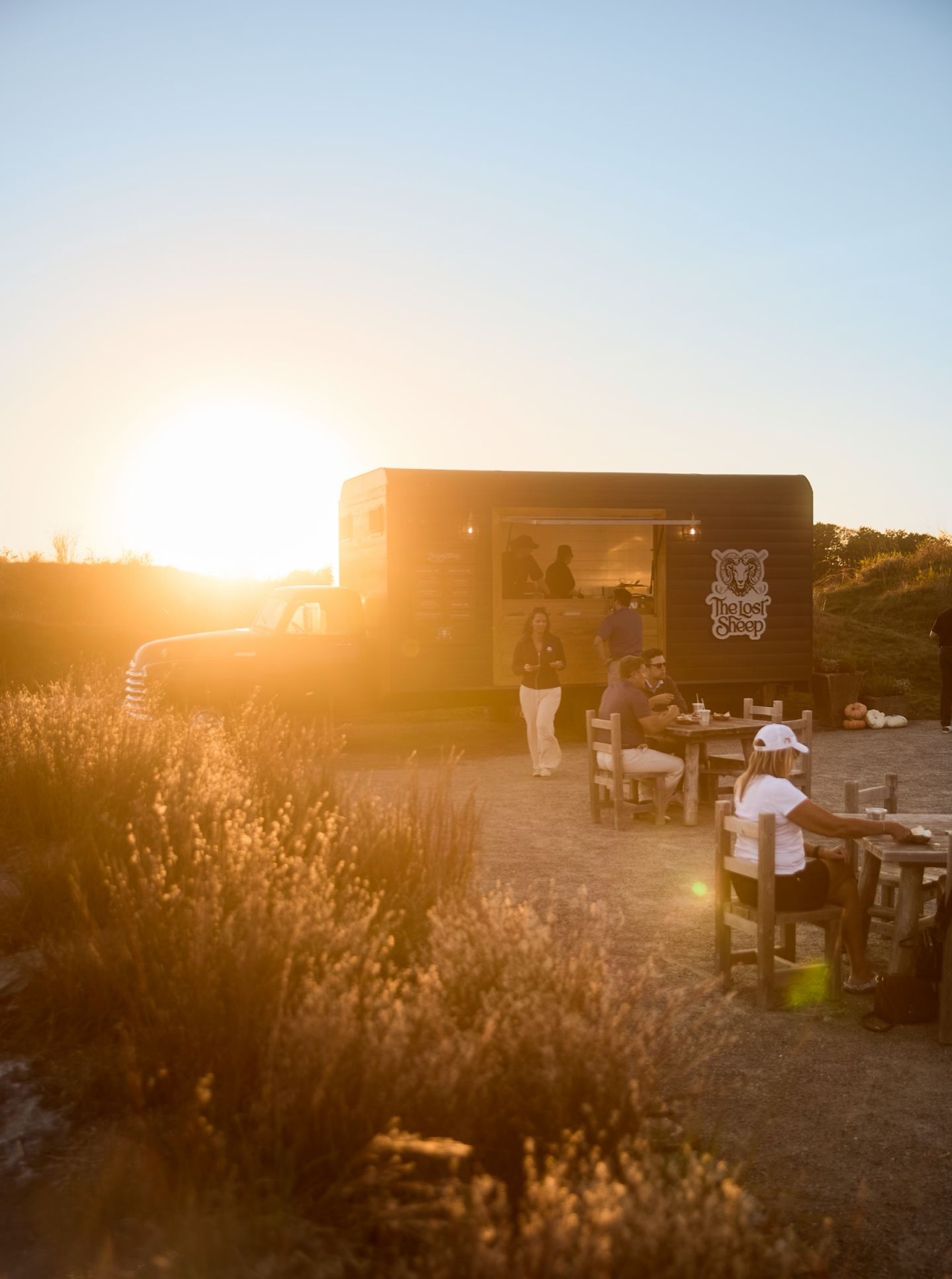 The Lost Sheep food truck backlit by a low sun, its serving window open with two staff inside, guests seated at wooden tables in front and tall grasses in the foreground.