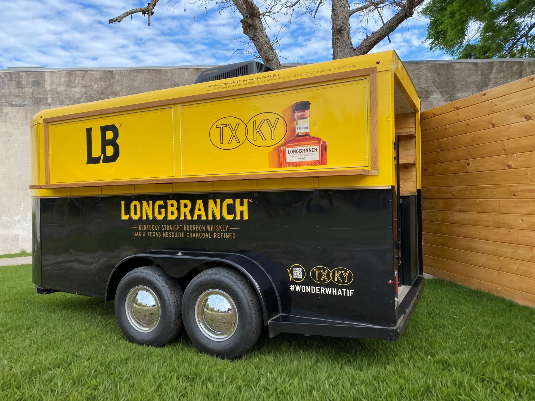 The Longbranch horse trailer closed up for the road, seen from the rear quarter, with the LB monogram, a TX KY roundel, a bottle graphic and the hashtag #WONDERWHATIF on its yellow and black sides.