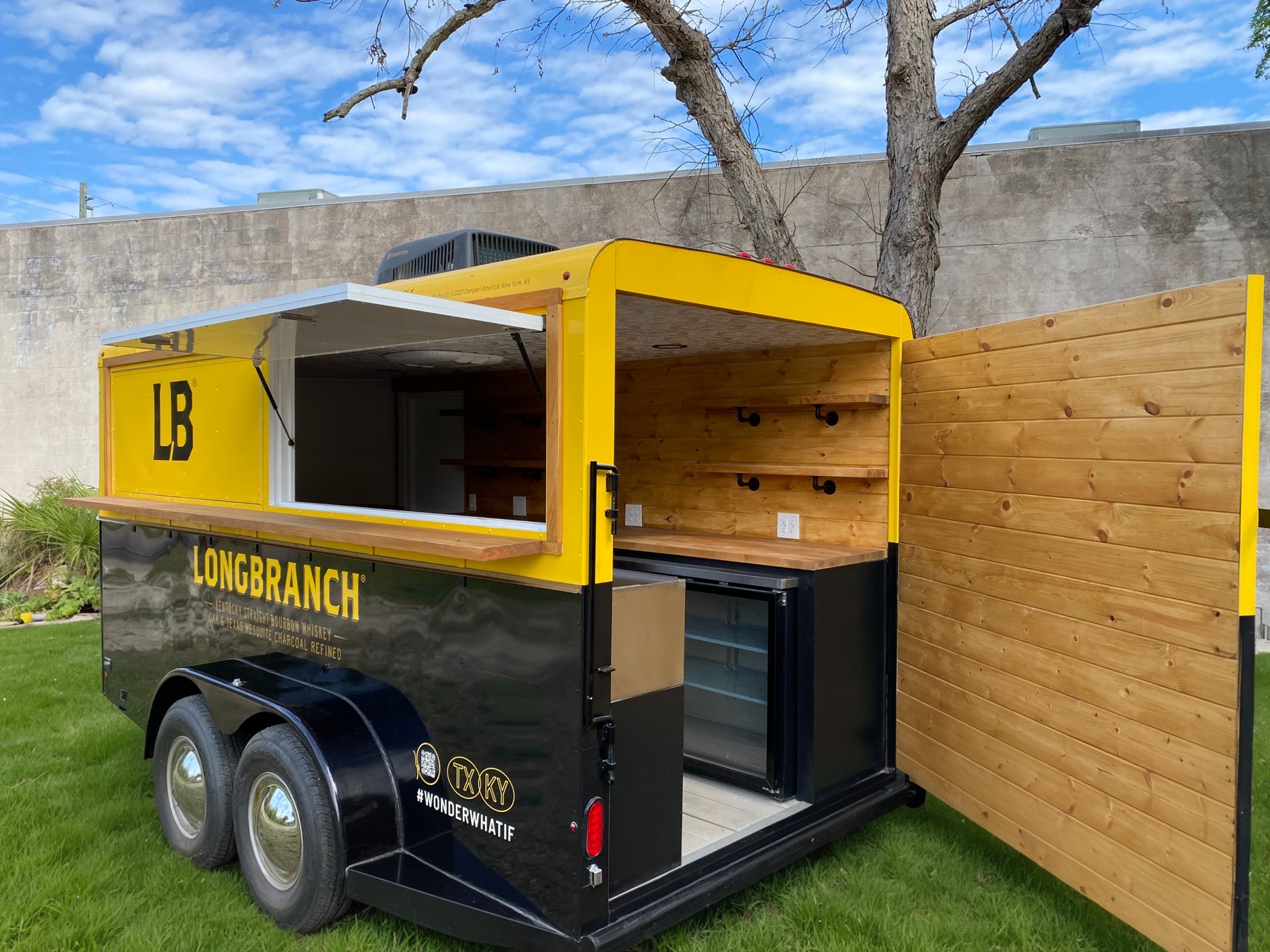 A yellow-and-black Longbranch horse trailer parked on grass, its side hatch propped open over a wooden counter and its rear door swung wide to show a knotty-pine bar with wooden shelves and an under-counter glass-door refrigerator.