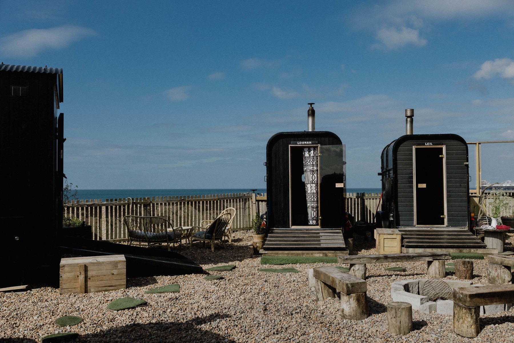Two small black wooden saunas with chimneys stand on a gravel site behind a fence at the edge of the sea, with log seats in the foreground.