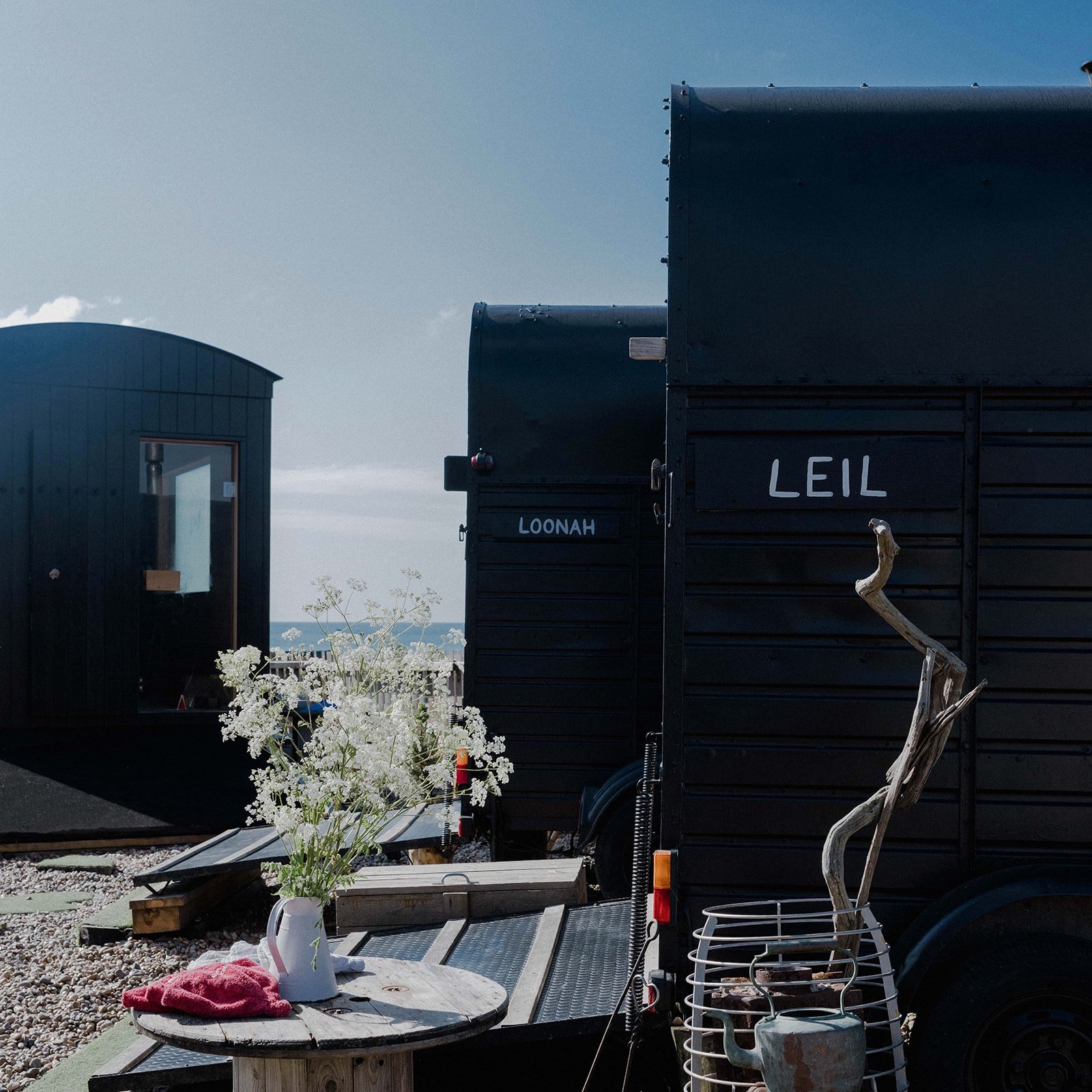 The black converted horsebox sauna Leil, its name painted on the side, beside a second horsebox and a wooden sauna on the shingle at Brighton, with a jug of wildflowers on a cable-reel table.
