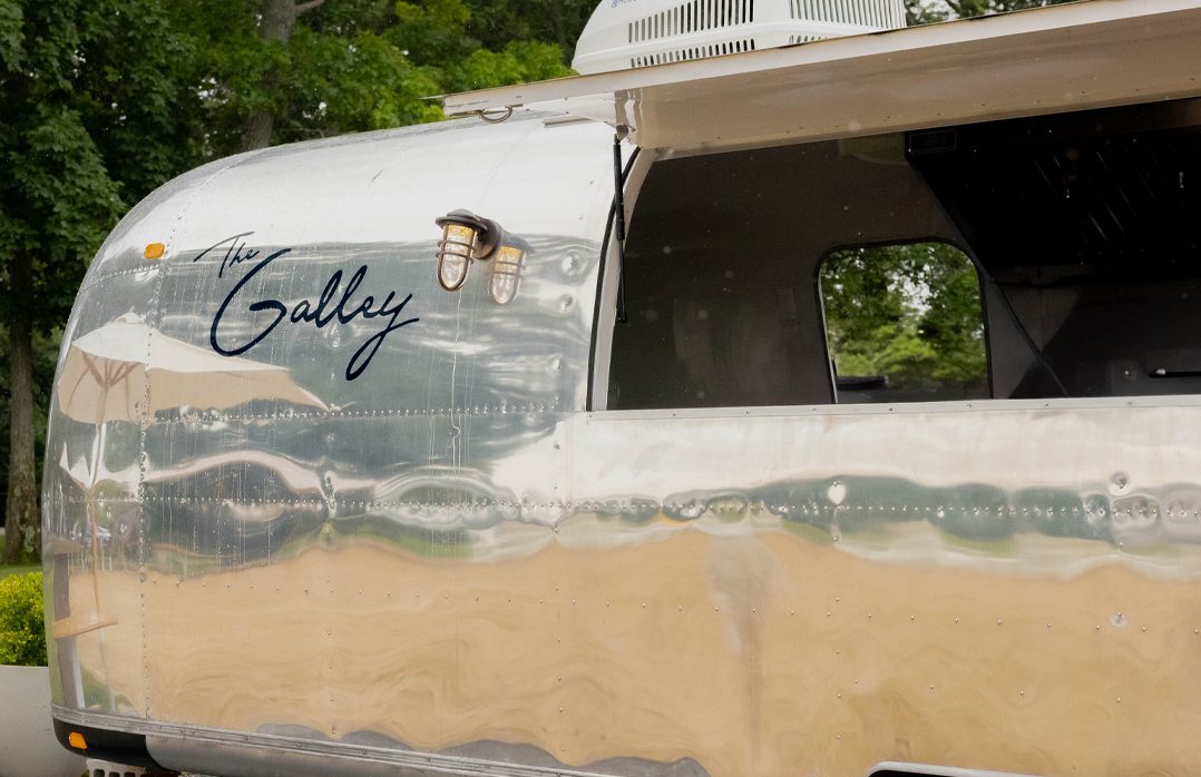 Close view of the Airstream's polished side with "The Galley" in navy script, two brass caged lamps and the raised serving hatch, trees reflected in the aluminum.