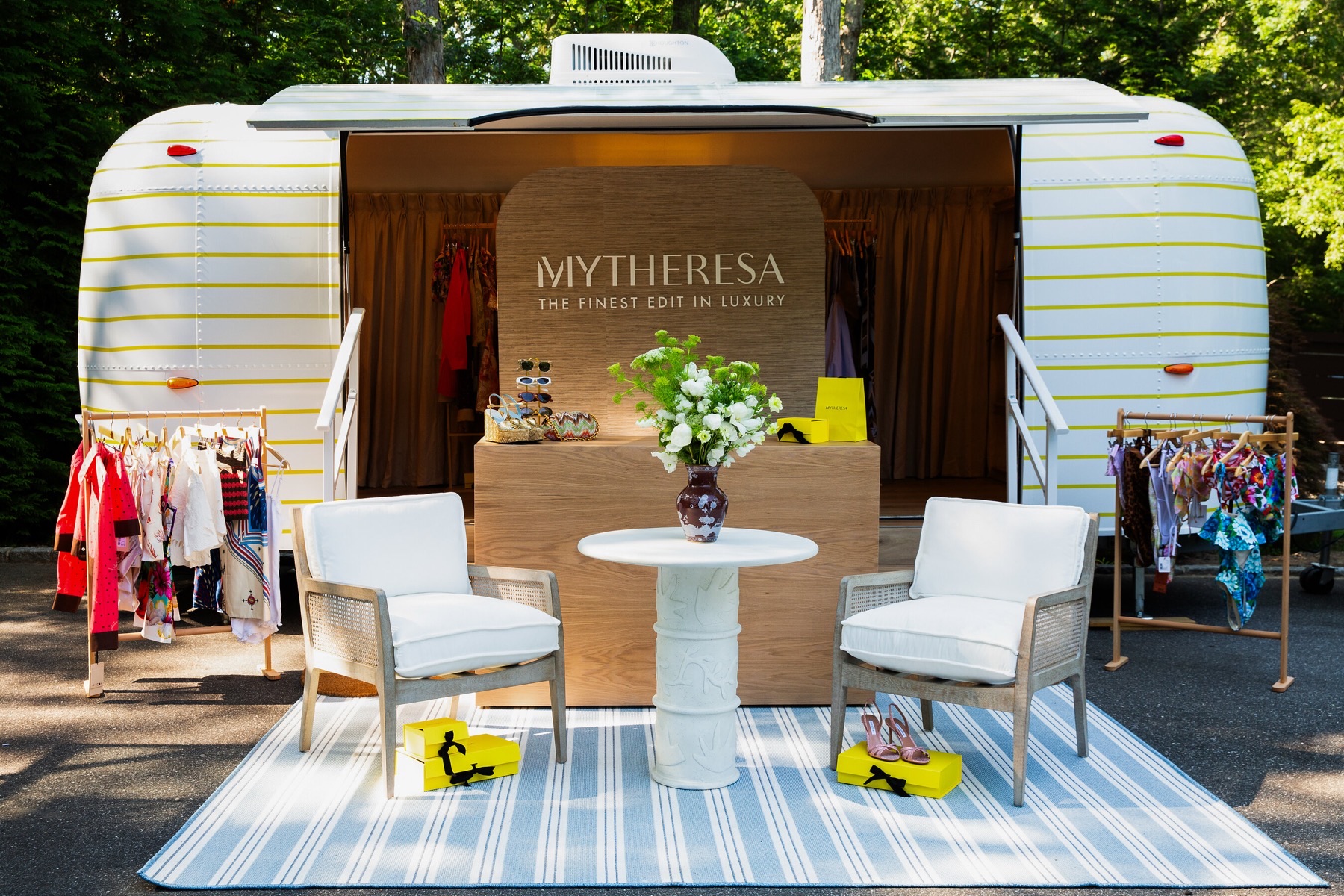 A polished Airstream trailer with white-and-yellow striped livery, its side opened to show a Mytheresa-branded counter and clothing racks, with two armchairs, a white side table with flowers and a striped rug set out in front.