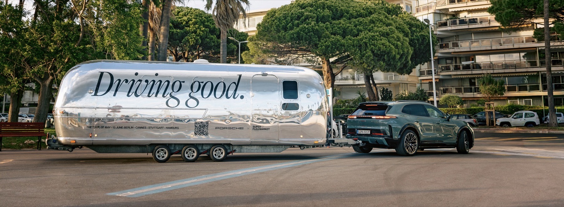 A polished aluminum Airstream trailer lettered 'Driving good.' with Porsche and Super Flea Market logos, hitched to a gray Porsche Cayenne on a palm- and pine-lined street.