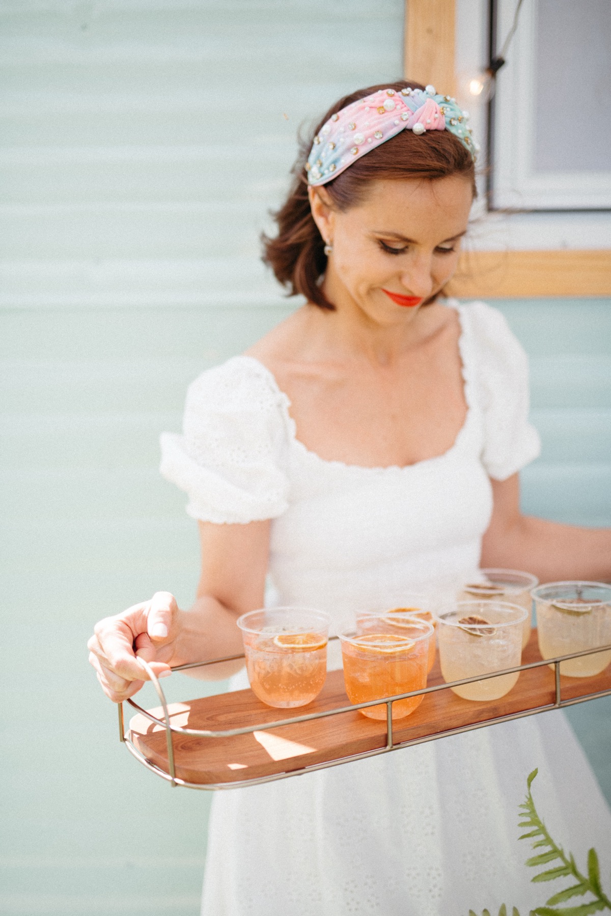 A woman in a white dress carries a wood-and-brass tray of iced drinks garnished with dried orange slices beside the camper's mint siding.