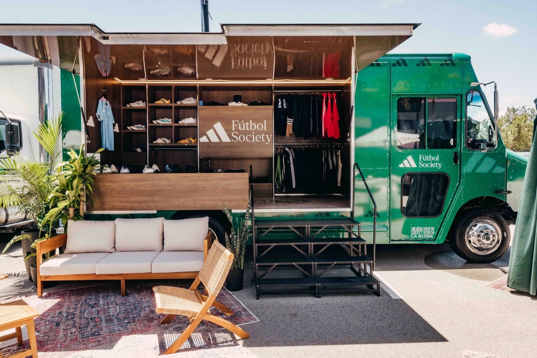 A green Adidas FĂștbol Society truck with its side opened to show wood-paneled shelving of sneakers and jerseys, a sofa, chair and rug set out in front.