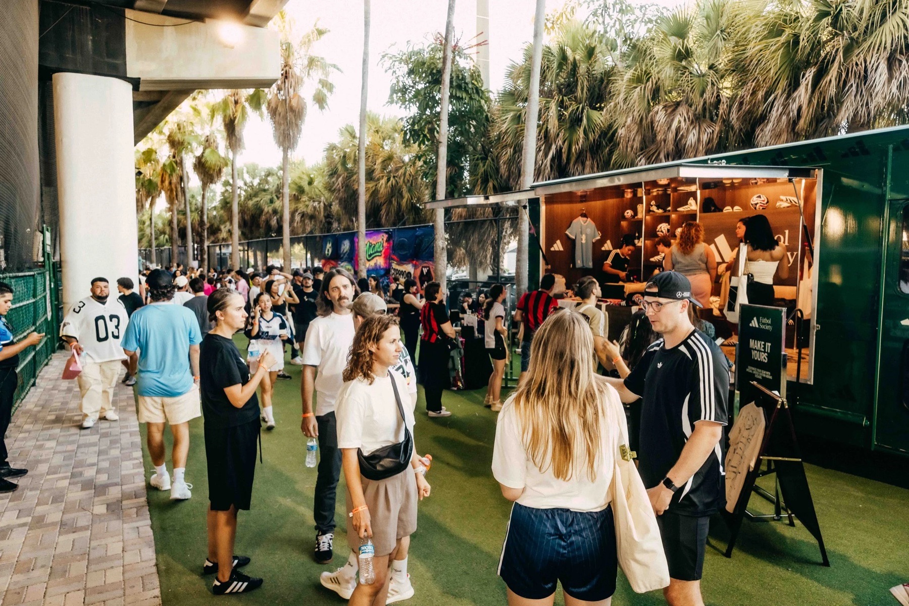 Visitors on artificial turf beside the open green Adidas truck at an evening event under palm trees, a 'Make it yours' sign by the counter.