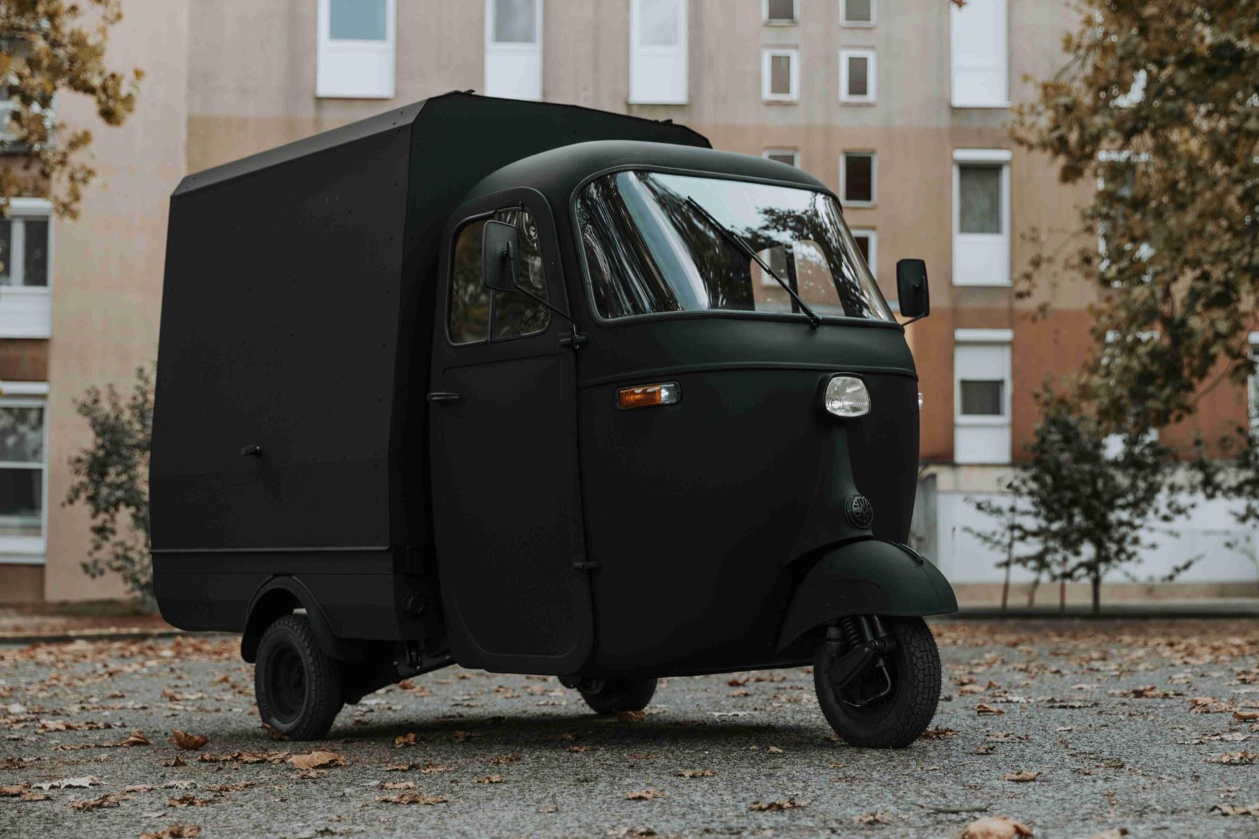 A matte black Piaggio Ape with a boxy closed rear body, parked on a gravel lot strewn with autumn leaves in front of an apartment building.