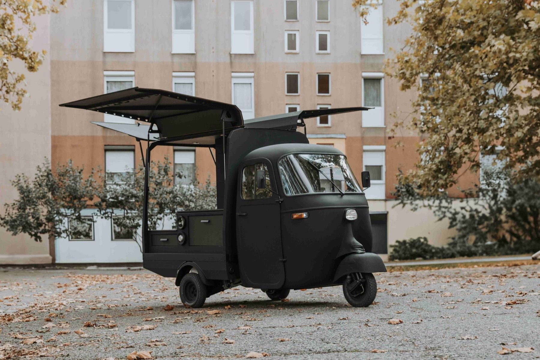 A matte black Piaggio Ape parked in front of an apartment building, with both gullwing roof panels raised over the service area.
