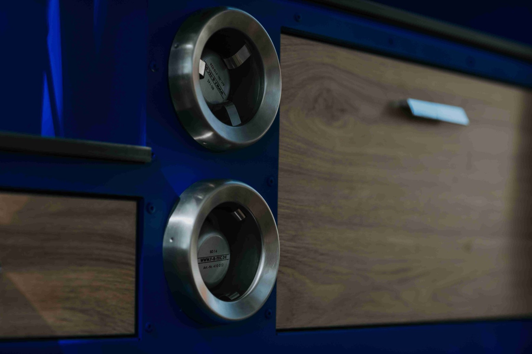 Close-up of two round stainless-steel cup dispensers set flush into the blue body of the Ape, between wood-faced drawer panels.
