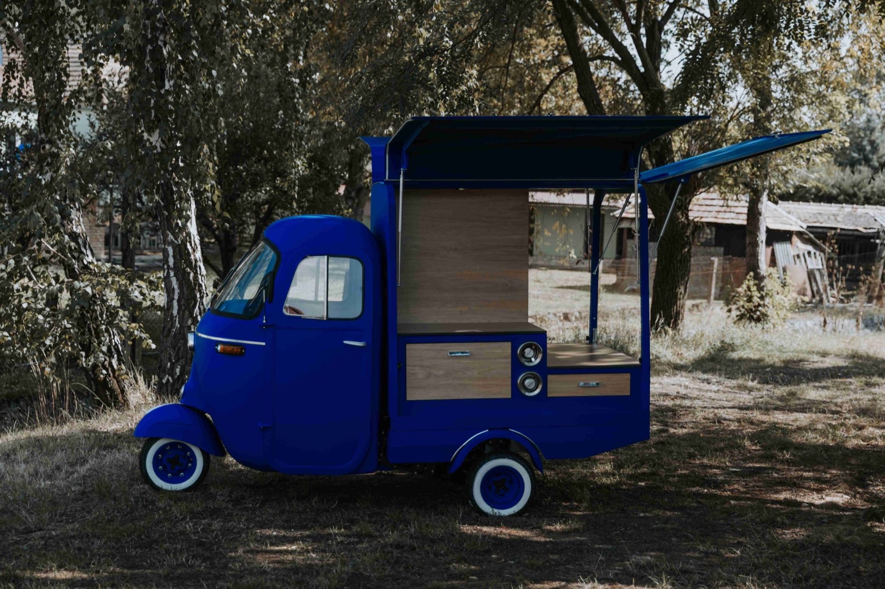 A bright blue three-wheeled Piaggio Ape on whitewall tires, parked on grass under trees with both gullwing roof panels raised over a wood-paneled service counter.